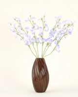 Un vase marron nervuré contient le bouquet de fleurs artificielles en soie, pois lavande, avec des pétales délicats et des tiges vertes sur un fond blanc cassé uni pour une atmosphère subtile de cottage.
