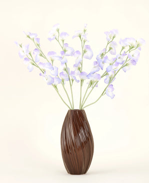 Un vase marron nervuré contient le bouquet de fleurs artificielles en soie, pois lavande, avec des pétales délicats et des tiges vertes sur un fond blanc cassé uni pour une atmosphère subtile de cottage.