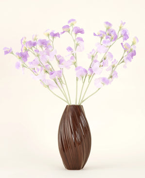 Un vase marron texturé contient le bouquet de fleurs artificielles en soie, des fleurs en soie violet-violet clair avec des tiges verdâtres sur un fond uni et clair.