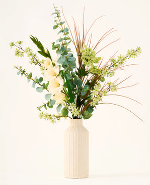 Dans un vase beige se trouve le bouquet "Wild Ivory" de fleurs artificielles en soie, avec des feuilles vertes, de l'eucalyptus, des fleurs blanches, de petites fleurs vertes et de longues et fines herbes brunes sur un fond clair.