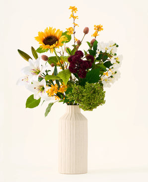 Dans un vase de couleur crème se trouve le bouquet de fleurs artificielles en soie 'Mom's Garden', un arrangement de tournesols, de lys, de fleurs blanches et bordeaux et de feuilles vertes sur un fond uni - le faisant ressembler à un bouquet de fleurs artificielles rappelant une bordure de jardin d'été.