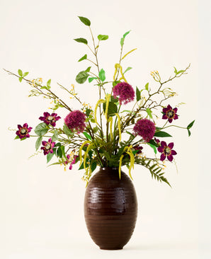 Dans un vase marron nervuré se trouve le bouquet "Woodland Charm" de fleurs artificielles en soie, avec des alliums violets, des fleurs rose foncé, des brindilles jaunes et des feuilles vertes sur un fond uni et clair.