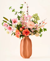 Dans un vase en terre cuite se trouve le bouquet coloré 'Yasmine's Choice' de fleurs artificielles en soie, composé de lys roses, de roses pêche et de feuilles vertes, composé de fleurs en soie de haute qualité d'Easyplants, le tout magnifiquement disposé sur un fond clair.