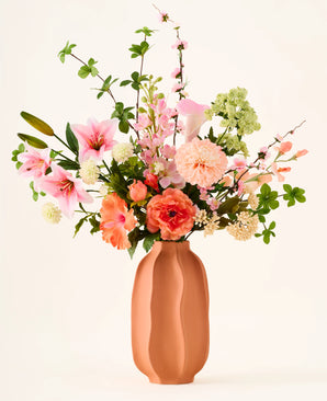 Dans un vase en terre cuite se trouve le bouquet coloré 'Yasmine's Choice' de fleurs artificielles en soie, composé de lys roses, de roses pêche et de feuilles vertes, composé de fleurs en soie de haute qualité d'Easyplants, le tout magnifiquement disposé sur un fond clair.
