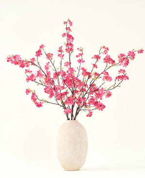 Le bouquet de fleurs artificielles en soie Fleur de cerisier fuchsia 150 cm présente des tiges de fleurs de cerisier fuchsia vibrantes avec des feuilles vertes dans un simple vase blanc, qui complètera n'importe quel intérieur sur un fond clair.