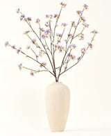 Un grand vase en céramique de couleur crème contient un certain nombre de fines branches avec le texte Bouquet de fleurs artificielles en soie Branche de fleurs artificielles violette, sur un fond uni et clair.