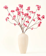Le bouquet de fleurs artificielles en soie Fleur de cerisier fuchsia 114 cm ajoute une touche de couleur vibrante à votre intérieur avec ses branches saisissantes et ses fleurs fuchsia, joliment présentées dans un vase en céramique beige sur un fond uni blanc cassé.