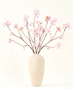Le bouquet de fleurs artificielles en soie Fleur de cerisier rose 114 cm a des branches longues et fines avec des bouquets de fleurs rose clair dans un vase en céramique beige, qui forment un contraste saisissant sur le fond blanc uni.