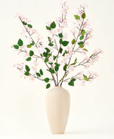 Un vase en céramique beige contient plusieurs longues branches avec le bouquet de fleurs artificielles en soie de feuilles roses et vertes de fleurs de pêcher, sur un fond blanc uni - parfait pour un intérieur floral artificiel.