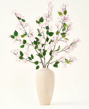 Un vase en céramique beige contient plusieurs longues branches avec le bouquet de fleurs artificielles en soie de feuilles roses et vertes de fleurs de pêcher, sur un fond blanc uni - parfait pour un intérieur floral artificiel.