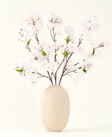 Le bouquet de fleurs artificielles en soie Cherry Blossom Blush 119 cm présente de délicates fleurs de cerisier rose clair avec des feuilles vertes dans un vase beige, idéal comme décoration de fleurs artificielles sur un fond clair.
