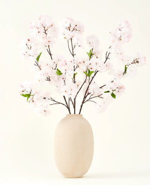 Le bouquet de fleurs artificielles en soie Cherry Blossom Blush 119 cm présente de délicates fleurs de cerisier rose clair avec des feuilles vertes dans un vase beige, idéal comme décoration de fleurs artificielles sur un fond clair.