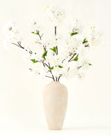 Le bouquet de fleurs artificielles en soie Fleur de cerisier crème 119 cm présente d'élégantes branches marron avec de fausses fleurs blanches douces et des feuilles vertes, présentées dans un simple vase beige sur un fond clair uni.