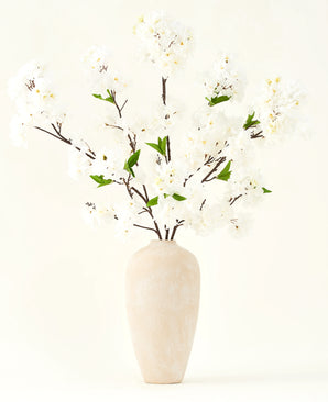 Le bouquet de fleurs artificielles en soie Fleur de cerisier crème 119 cm présente d'élégantes branches marron avec de fausses fleurs blanches douces et des feuilles vertes, présentées dans un simple vase beige sur un fond clair uni.