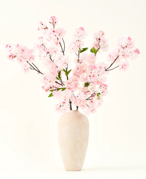 De Zijden kunstbloemen boeket Kersenbloesem roze 119 cm, gerangschikt in een beige keramische vaas met delicate groene bladeren, creëert een sereen interieuraccent tegen een eenvoudige lichte achtergrond.
