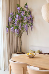 Une plante artificielle de glycine mesurant 180 cm avec des grappes de fleurs bleu lilas se trouve près d'une fenêtre aux rideaux beiges, à côté d'une table en bois avec un bol de citrons et un banc avec des coussins verts.