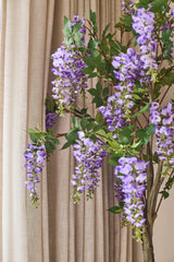 Une plante artificielle Wisteria de 150 cm aux feuilles vertes est suspendue devant les rideaux beiges.