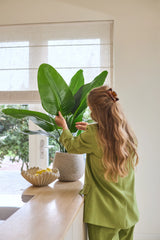 Une femme aux longs cheveux ondulés, vêtue d'un costume vert, s'occupe d'une plante artificielle Strelitzia Real Touch Deluxe de 90 cm posée sur un comptoir à côté d'un bol de citrons dans une pièce lumineuse avec une fenêtre.