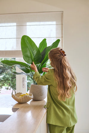 Une femme aux longs cheveux ondulés, vêtue d'un costume vert, s'occupe d'une plante artificielle Strelitzia Real Touch Deluxe de 90 cm posée sur un comptoir à côté d'un bol de citrons dans une pièce lumineuse avec une fenêtre.