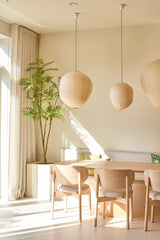 Une salle à manger lumineuse et minimaliste est meublée de chaises en bois clair, d'une table, de trois lampes suspendues en papier, de la plante artificielle Mahonia de 210 cm en pot et de rideaux allant du sol au plafond près d'une grande fenêtre laissant entrer la lumière du soleil.