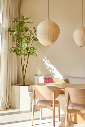 Un espace de vie lumineux et minimaliste avec des chaises en bois, un canapé blanc aux coussins colorés, de grandes fenêtres avec rideaux, deux lampes suspendues lanternes rondes en papier et la Plante Artificielle Mahonia de 210 cm, qui apporte une touche de vert. La lumière du soleil projette des ombres sur les murs.