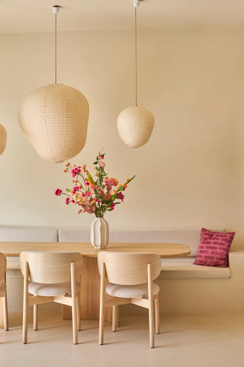 Een minimalistische eethoek met een tafel en stoelen van licht hout, beige muren, drie hanglampen in de vorm van papieren lantaarns, een vaas met het boeket ‘Wild Peach’ van Zijden kunstbloemen dat voor een levendig kleuraccent zorgt, en een ingebouwde bank met daarop een decoratief roze kussen.