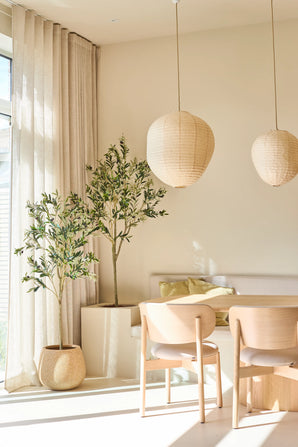 Une salle à manger lumineuse et minimaliste est meublée de deux chaises en bois clair, d'une petite table, de lanternes en papier et de l'olivier artificiel de 180 cm, qui ajoute une touche de style méditerranéen. Des arbres d'intérieur en pot, des rideaux beiges translucides et la lumière du soleil pénétrant à travers les grandes fenêtres complètent cet espace de détente.