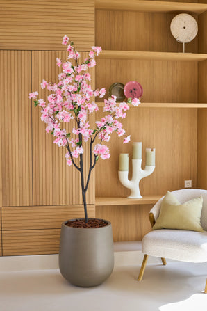 Une fleur de cerisier artificielle de 180 cm rose, un arbre en pot aux fleurs roses, se dresse à côté d'une chaise blanche avec des coussins dans une pièce moderne avec des étagères en bois et des objets décoratifs.