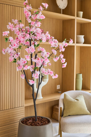 La Fleur de Cerisier Artificielle 180 cm rose, avec ses délicates fleurs roses, trône dans un salon moderne avec des étagères en bois, des céramiques, un fauteuil beige avec un coussin vert et une décoration minimaliste.