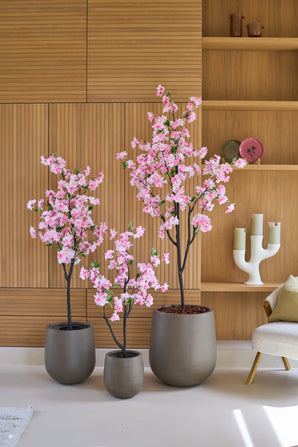 Trois cerisiers artificiels de 180 cm dans un pot rose, avec des fleurs roses, fabriqués à partir de fleurs de cerisier en soie de haute qualité, se dressent devant un mur en bois avec des étagères intégrées, à côté d'une chaise crème et d'objets de décoration.