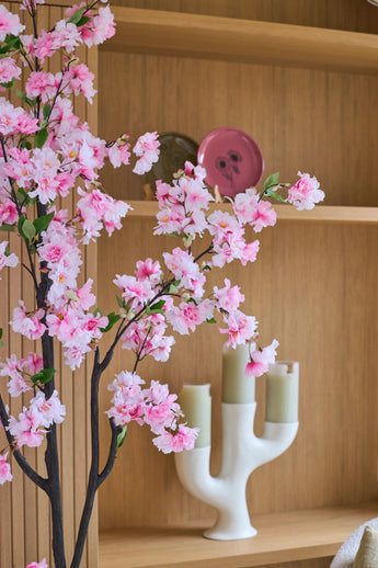 Une fleur de cerisier artificielle de 180 cm de couleur rose se dresse devant des étagères en bois sur lesquelles sont disposés une assiette rose, un bol vert et un chandelier blanc avec trois bougies vert clair.