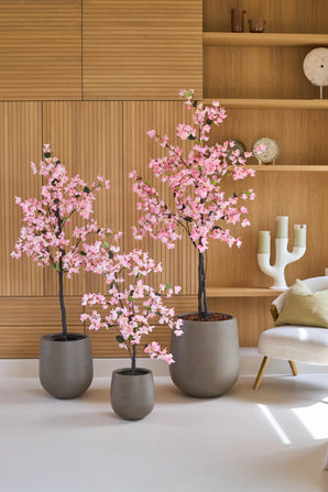 Trois bougainvilliers artificiels de 180 cm, de couleur rose et avec des fleurs roses, se dressent devant des étagères murales en bois, à côté d'une chaise blanche moderne avec un coussin.