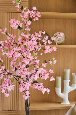 Un bougainvillier artificiel de 180 cm, de couleur rose avec des fleurs roses, se dresse devant des étagères en bois sur lesquelles se trouvent une assiette décorative et un chandelier blanc avec trois bougies vertes.