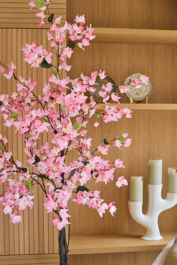Un bougainvillier artificiel de 180 cm, de couleur rose avec des fleurs roses, se dresse devant des étagères en bois sur lesquelles se trouvent une assiette décorative et un chandelier blanc avec trois bougies vertes.