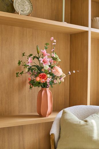 Sur une étagère en bois à côté d'une chaise confortable avec un coussin vert se trouve un vase rose avec le bouquet de fleurs artificielles en soie « Yasmine's Choice », créant une image intérieure chaleureuse et invitante.