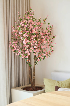 La plante de cerisier artificielle 120 cm rose, avec ses fleurs de cerisier roses, se trouve à côté d'une table en bois dans une pièce lumineuse et moderne, avec un rideau beige et un canapé clair derrière.