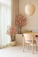 Une chambre minimaliste avec des meubles en bois clair, des rideaux de couleur crème et deux fleurs de cerisier artificielles de 150 cm de couleur rose, qui fournissent des fleurs roses. Une lanterne ronde en papier est suspendue au-dessus de la table à manger, avec une chaise rembourrée et un banc intégré.