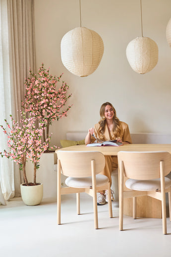 Une femme aux longs cheveux blonds est assise à une table en bois clair, lisant un livre et souriant. À côté d'elle se trouve la plante de cerisier artificielle de 150 cm rose, dont les fleurs roses apportent un accent vivant. Les lampes suspendues et la décoration minimaliste créent une atmosphère légère et cosy.