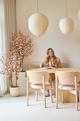 Une femme est assise, souriante, à une table avec un livre ouvert. La pièce est décorée avec des meubles en bois clair, des lanternes en papier, une douce lumière naturelle pénétrant à travers de hauts rideaux et une plante de cerisier artificielle de 120 cm rose qui ajoute une touche de fleur rose.