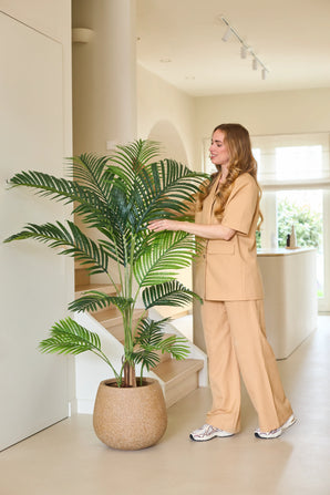 Une femme vêtue d'une robe beige se tient à l'intérieur et sourit en touchant les feuilles luxuriantes de la plante artificielle Phoenix 160 cm, qui se trouve à côté d'un escalier. La lumière naturelle remplit l’espace moderne et minimaliste et souligne son aspect luxueux.