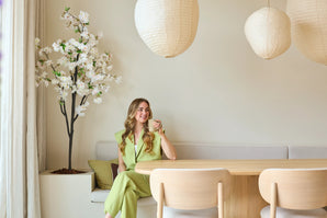 Une femme vêtue d'un costume vert clair est assise sur un banc près d'une table à manger en bois, avec une tasse à la main. Des lanternes décoratives en papier sont suspendues au-dessus d'elle et dans le coin se dresse une fleur de cerisier artificielle blanche de 180 cm, tandis qu'une douce lumière naturelle pénètre à travers une fenêtre.