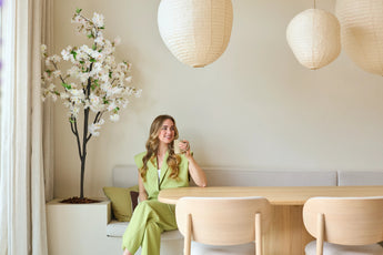 Une femme vêtue d'un costume vert clair est assise sur un banc près d'une table à manger en bois, avec une tasse à la main. Des lanternes décoratives en papier sont suspendues au-dessus d'elle et dans le coin se dresse une fleur de cerisier artificielle blanche de 180 cm, tandis qu'une douce lumière naturelle pénètre à travers une fenêtre.