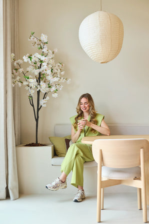 Une femme vêtue d'un costume vert clair est assise sur un banc, souriante et tenant une tasse à la main. A côté d'elle se trouve la Fleur de Cerisier Artificielle 180 cm blanche, placée dans une pièce moderne et minimaliste aux tons neutres et une lanterne ronde en papier au-dessus de sa tête.