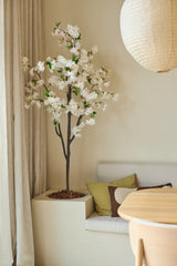 Une fleur de cerisier artificielle de 180 cm de couleur blanche côtoie un coin salon avec des coussins dans les tons vert et marron, près d'une table ronde en bois et d'une lanterne en papier dans une pièce à la lumière tamisée et aux tons neutres.