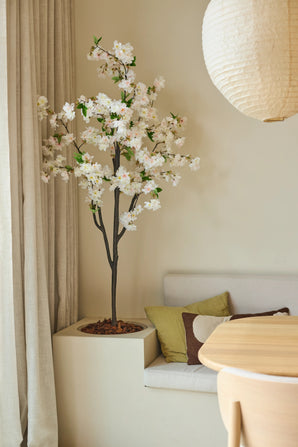 Une fleur de cerisier artificielle de 180 cm de couleur blanche côtoie un coin salon avec des coussins dans les tons vert et marron, près d'une table ronde en bois et d'une lanterne en papier dans une pièce à la lumière tamisée et aux tons neutres.
