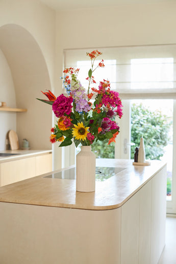 Un vase blanc avec le bouquet de fleurs artificielles en soie « Colourful Spirit » se dresse sur un îlot de cuisine en bois clair dans une cuisine lumineuse et moderne avec de grandes fenêtres et un décor neutre, créant un point focal durable et vivant.
