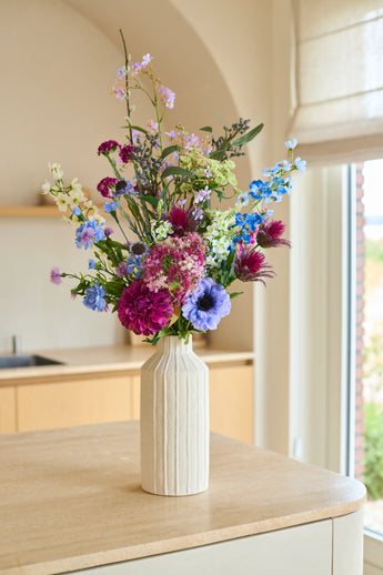 Le bouquet de fleurs artificielles en soie Purple Rain, un arrangement de fleurs en soie violettes, bleues, roses et blanches, est placé dans un grand vase en céramique de couleur crème. Ce bouquet fait main repose gracieusement sur un comptoir en bois clair, baigné d’une douce lumière naturelle – un véritable reflet du savoir-faire néerlandais.