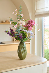 Sur un îlot de cuisine de couleur claire, près d'une fenêtre où pénètre la lumière naturelle, se trouve un vase vert avec des fleurs roses, violettes et blanches du bouquet de fleurs artificielles en soie 'Lavender Dreams'.