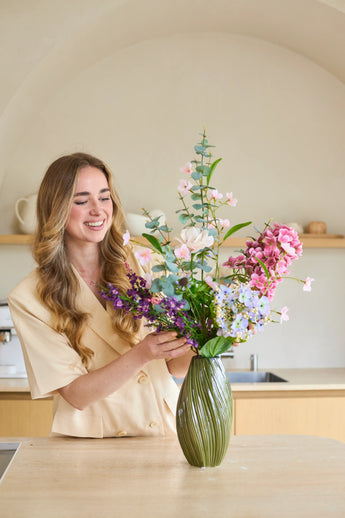 Une femme aux cheveux longs et ondulés dispose un bouquet de « Lavender Dreams » de fleurs artificielles en soie dans un vase vert dans sa cuisine moderne, souriante pendant qu'elle travaille. Le bouquet vibrant de fleurs en soie apporte une beauté intemporelle à sa maison.