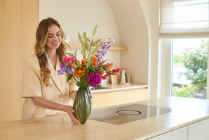 Une femme souriante arrangeant le bouquet « Fleurs artificielles en soie colorées Svenski » dans un vase vert sur un comptoir de couleur claire dans une cuisine au décor moderne et à la lumière naturelle.